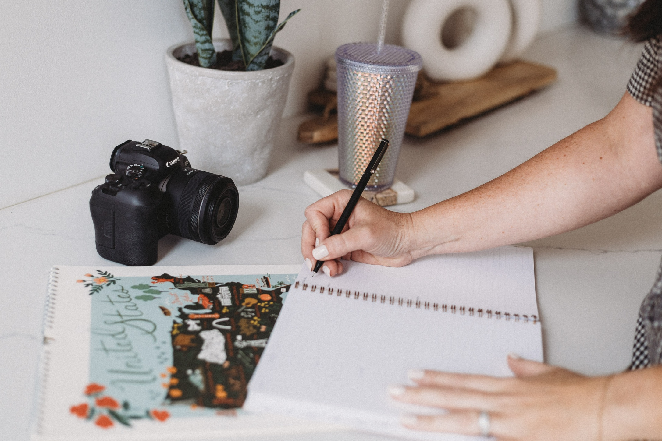 Hands writing in a notebook next to a camera on a white desk while planning content